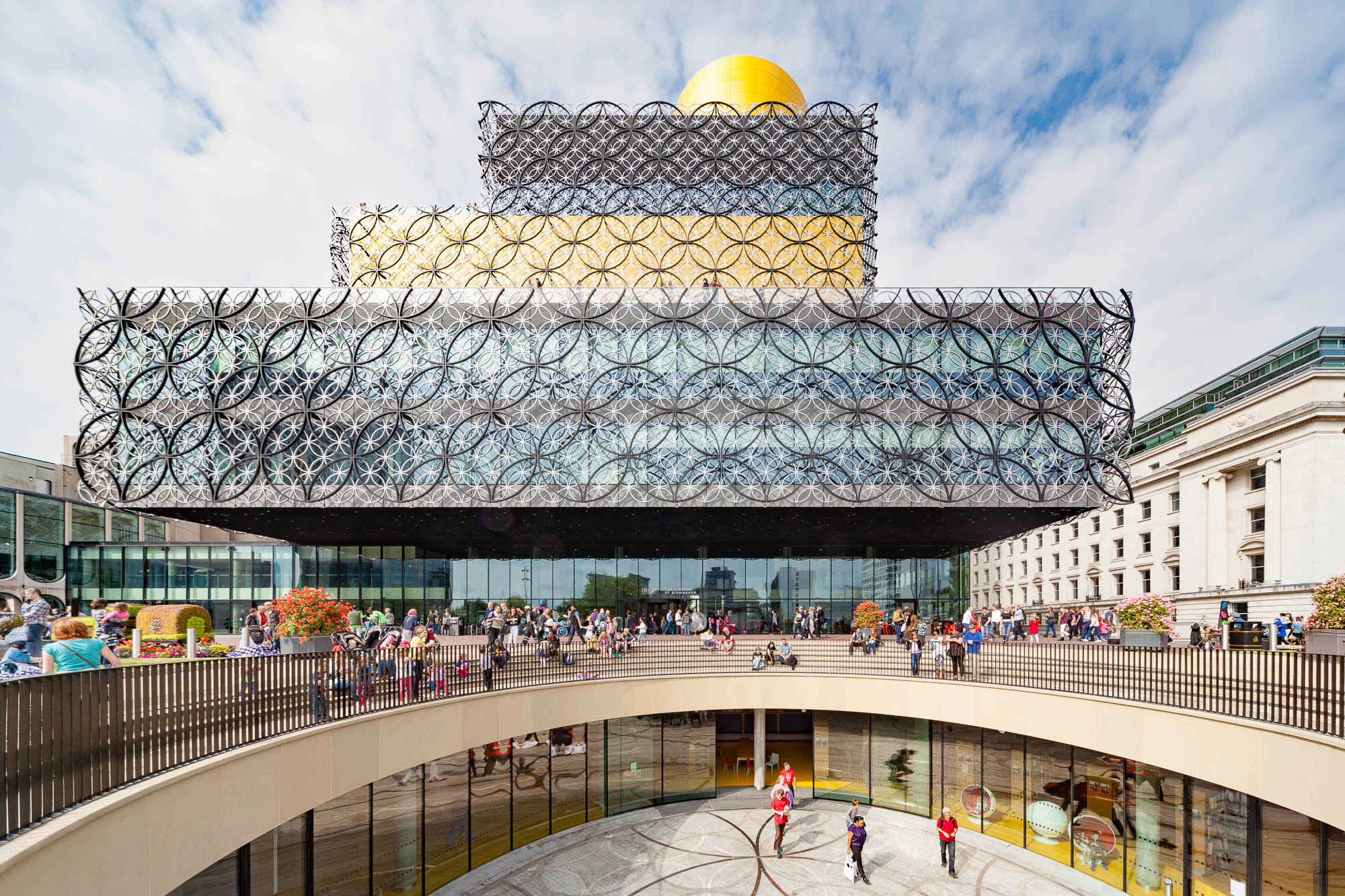 The image shows the modern Library of Birmingham with a gold-topped upper floor and decorative circular metalwork on its facade. People are gathered outside and on the entrance plaza under a partly cloudy sky.