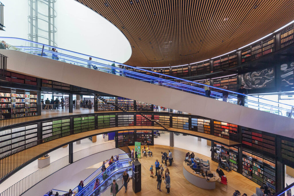 A modern, multi-level library interior with curved balconies, bookshelves lining the walls, people walking and reading, and a circular information desk on the wooden floor below.