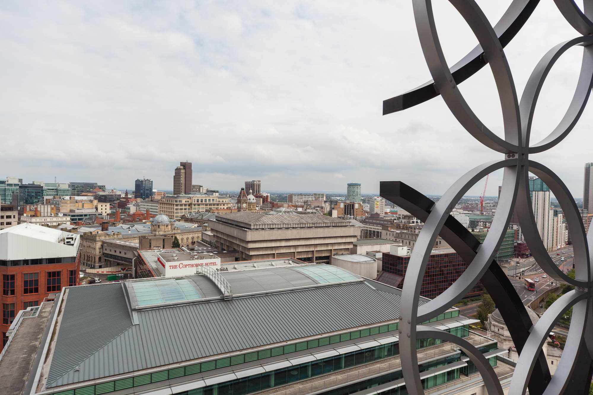 A cityscape view featuring modern and older buildings under a cloudy sky, with a decorative metal structure partially visible on the right side of the image.