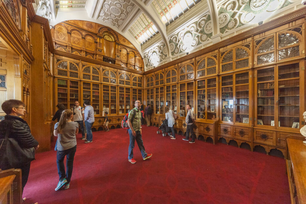 A group of people stand and walk inside an ornate library with tall wooden bookshelves, glass doors, and a high, decorative ceiling with stained glass panels. The floor is covered with red carpet.