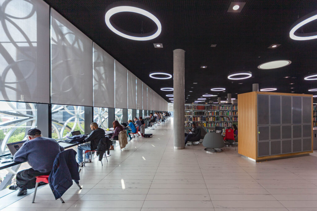 People are seated at desks by large windows in a modern library with circular lights on the ceiling, using laptops and reading. Rows of bookshelves are visible in the background.