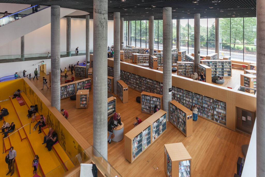 A modern library interior with tall concrete columns, large windows, wooden floors, bookshelves, and seating areas where people are reading or walking. The space is open, bright, and organized with natural light.
