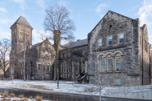 A large, historic stone building with arched windows and a tall clock tower stands beside a snowy sidewalk and leafless trees under a partly cloudy sky.
