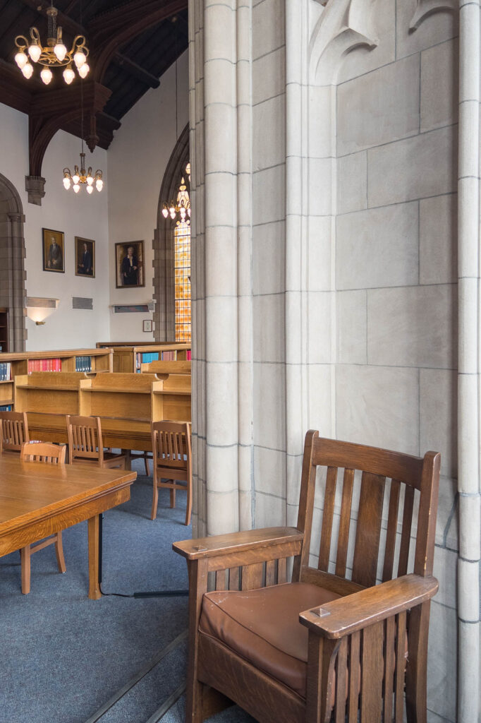 A wooden chair with a brown leather seat is placed against a stone wall in a library. Bookshelves, wooden tables, chairs, and framed portraits are visible in the background under hanging lights.