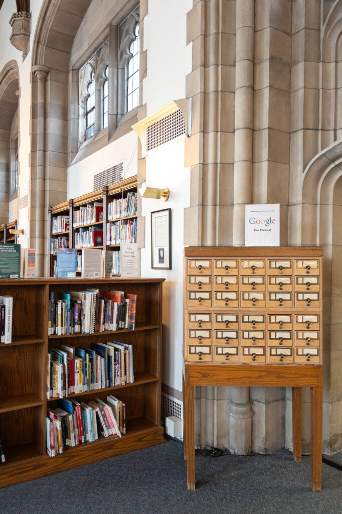 A wooden card catalog sits next to bookshelves filled with books in a library with arched windows and stone walls. A small sign on the card catalog reads, “Google: The first search engine.”.