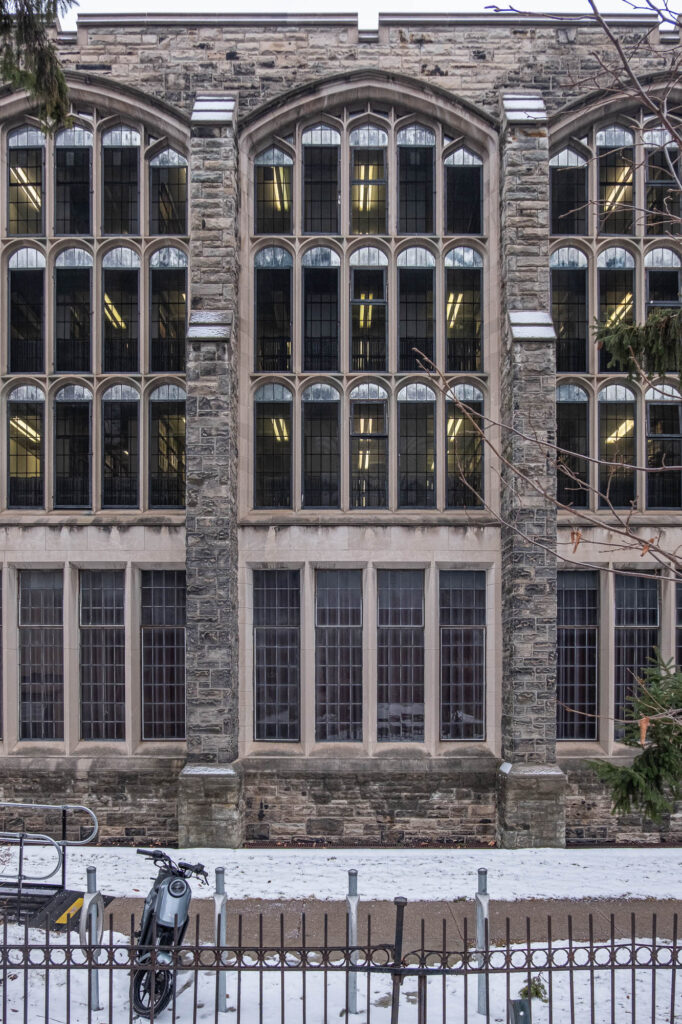 A stone building with large, multi-pane arched windows spanning three stories. Some windows have lights on inside. The ground is covered with snow, and a moped is parked near a metal fence in front of the building.