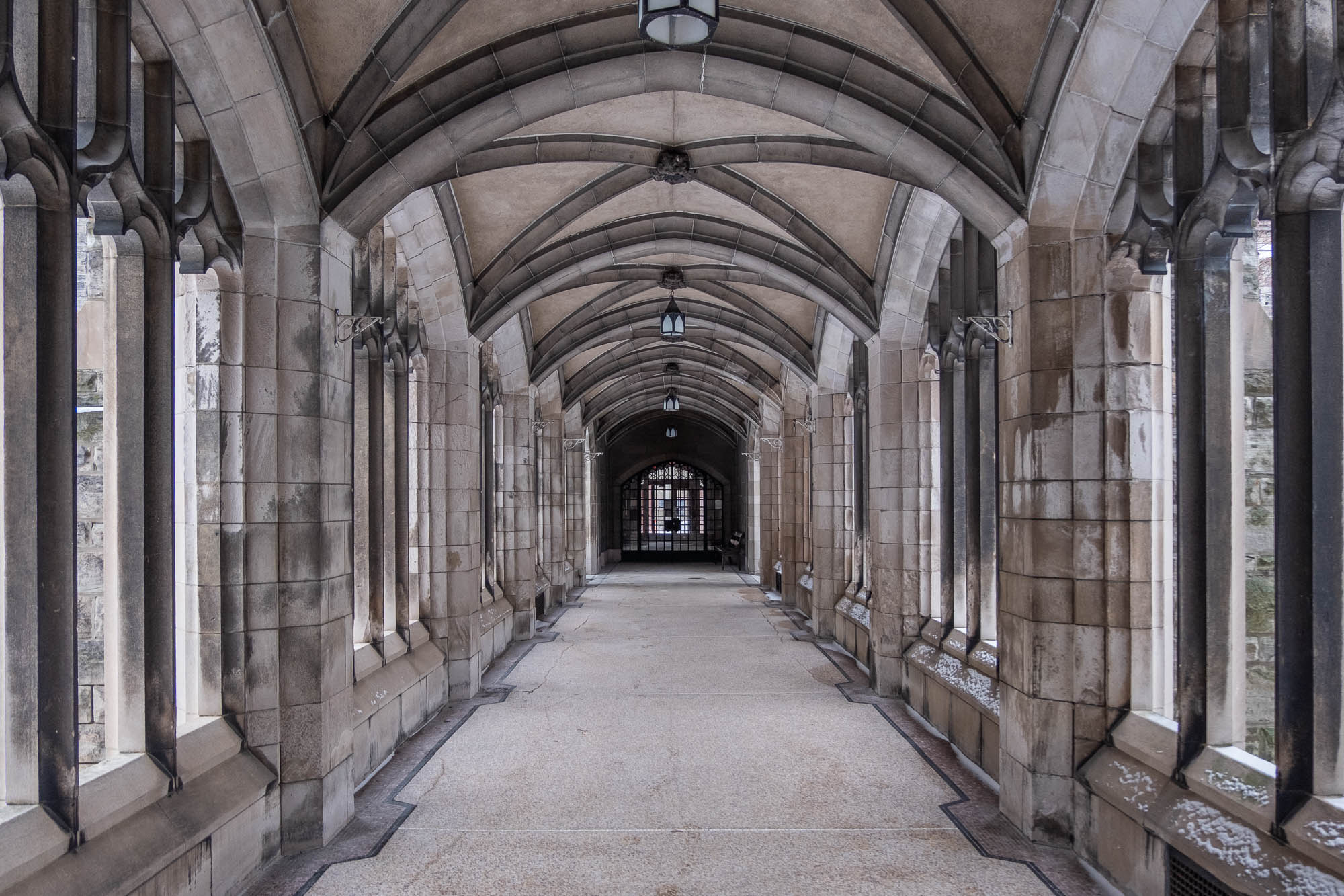 A stone hallway with arched ceilings and symmetrical columns on both sides, featuring gothic architectural details, leading to a gated entrance at the end.