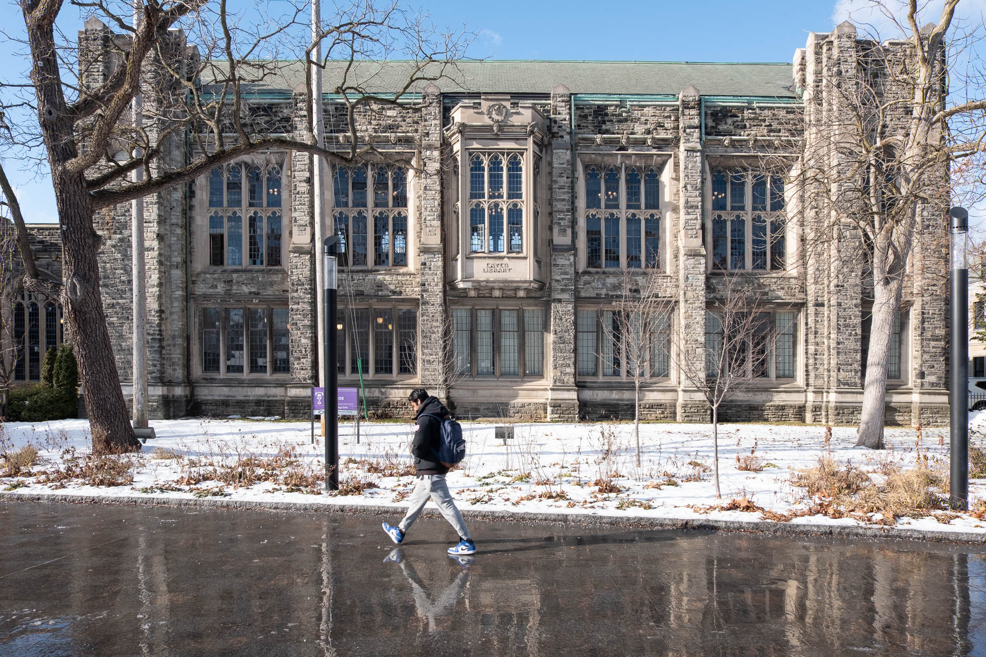 Exterior of a stone library building in Collegiate Gothic style. There is snow on the ground and the sky is clear. A person walks in the forefront.