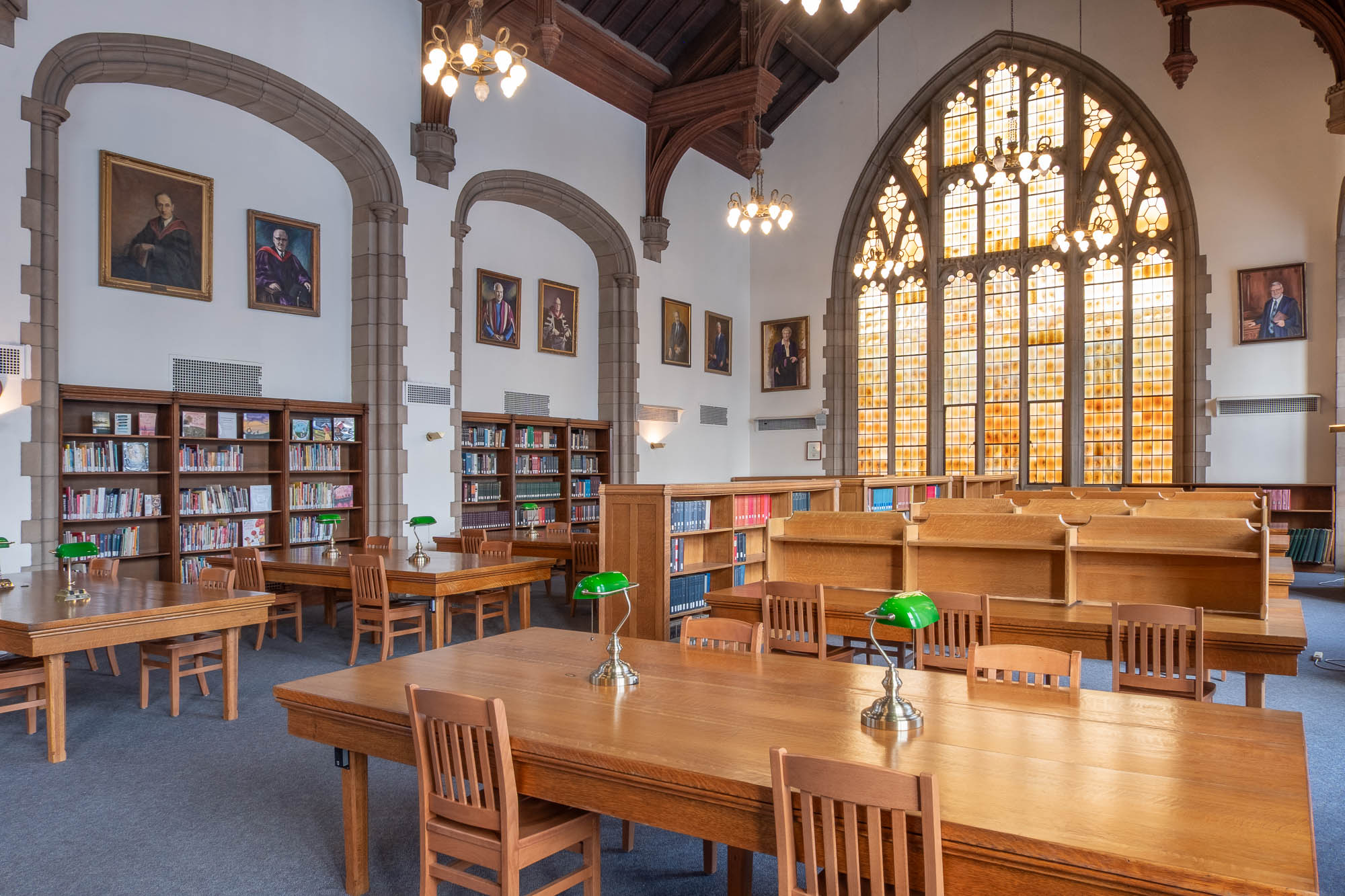 A spacious, traditional library room with high vaulted wooden ceilings, large arched windows, wooden tables and chairs, bookshelves, chandeliers, and framed portraits on the walls.