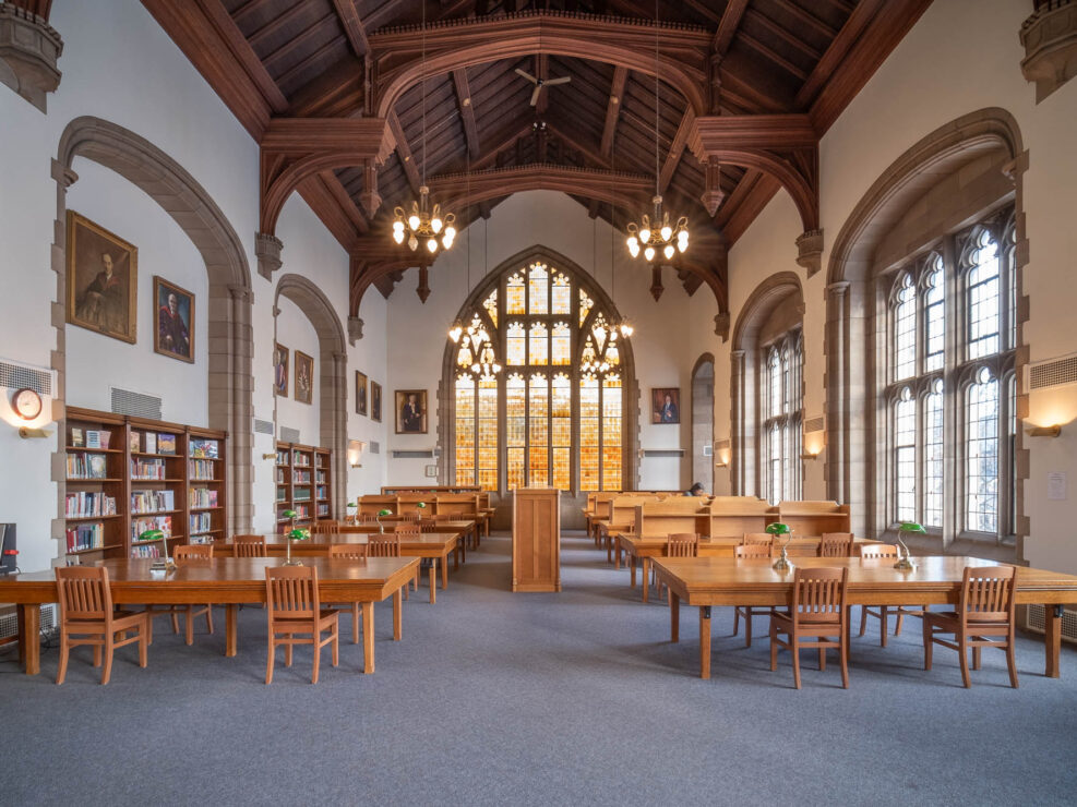A spacious, traditional library room with high vaulted wooden ceilings, large arched windows, wooden tables and chairs, bookshelves, chandeliers, and framed portraits on the walls.