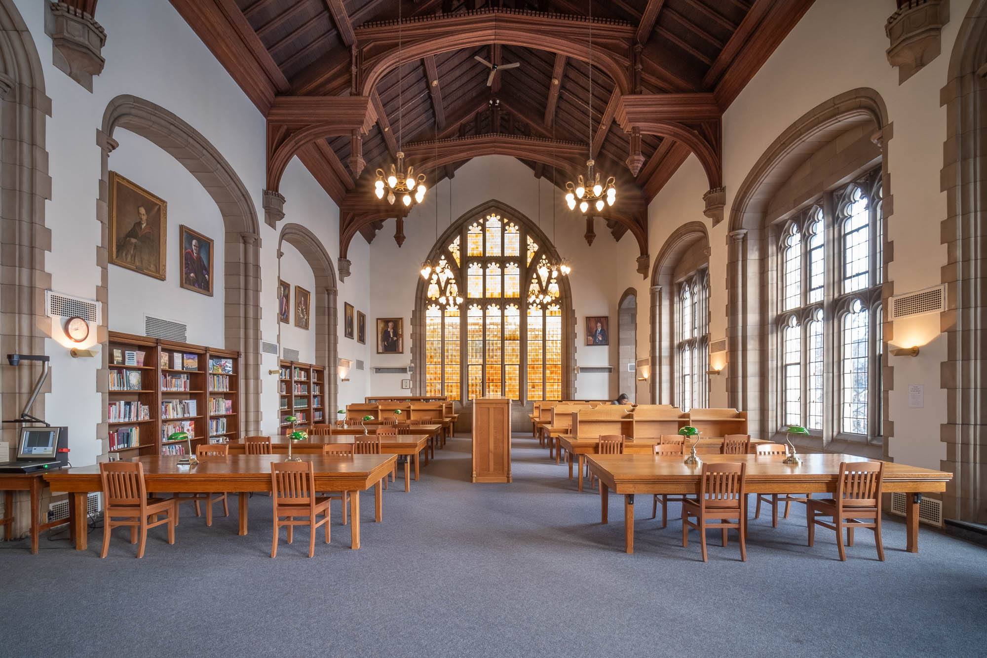 A spacious, traditional library room with high vaulted wooden ceilings, large arched windows, wooden tables and chairs, bookshelves, chandeliers, and framed portraits on the walls.