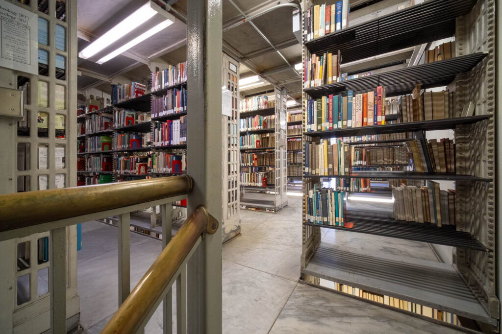 Metal bookshelves filled with books on multiple levels. In the forefront are brass railings.