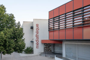 A modern building with white walls and red accents features the word médiathèque vertically on its facade. There are large windows with horizontal blinds and a tree partially visible on the left.