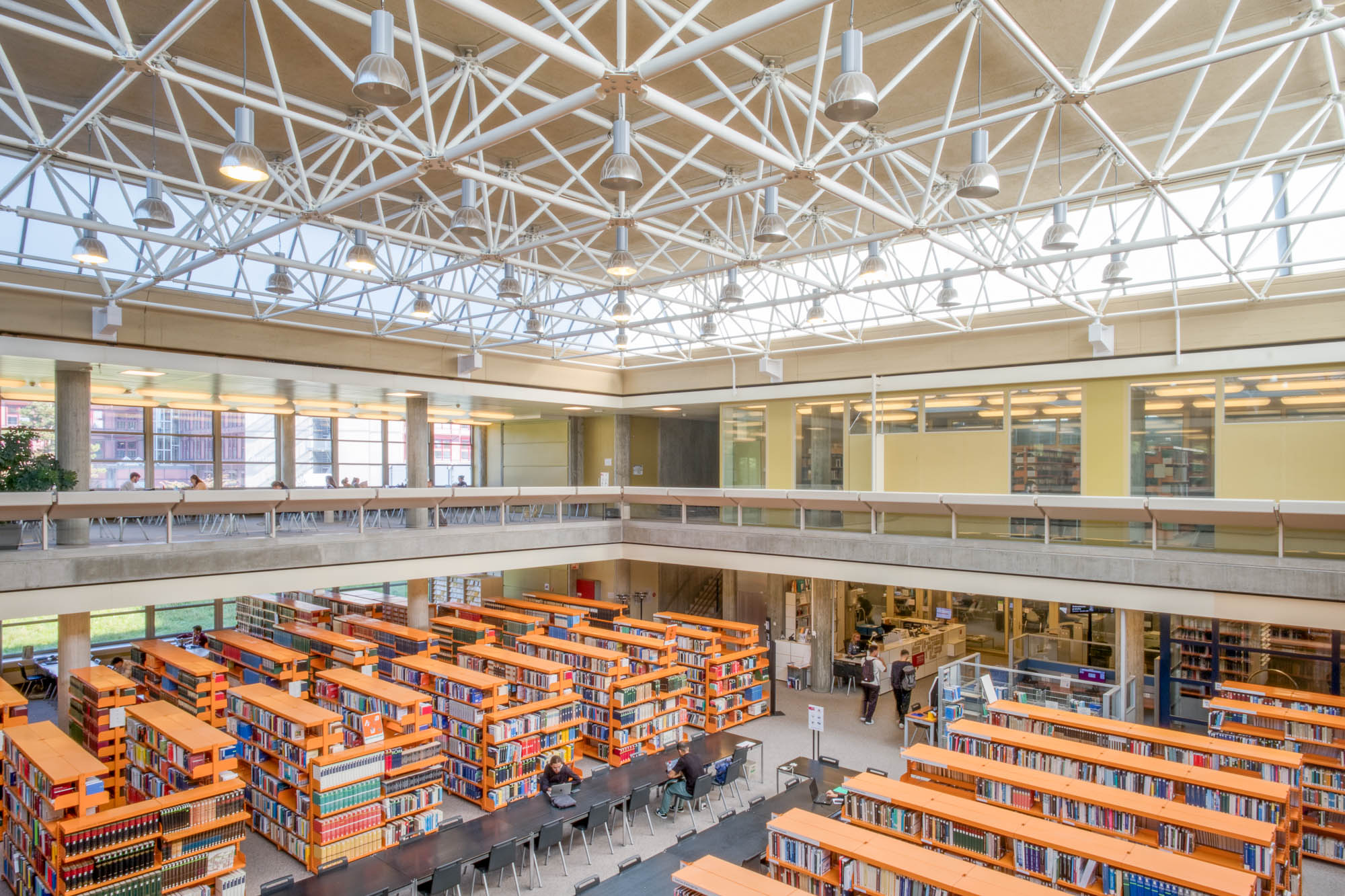 A spacious, modern library interior with tall bookshelves, tables and chairs arranged for study, a mezzanine level with glass railings, and a ceiling with exposed white beams and large windows letting in natural light.