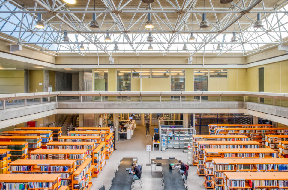 A spacious, modern library interior with tall bookshelves, tables and chairs arranged for study, a mezzanine level with glass railings, and a ceiling with exposed white beams and large windows letting in natural light.