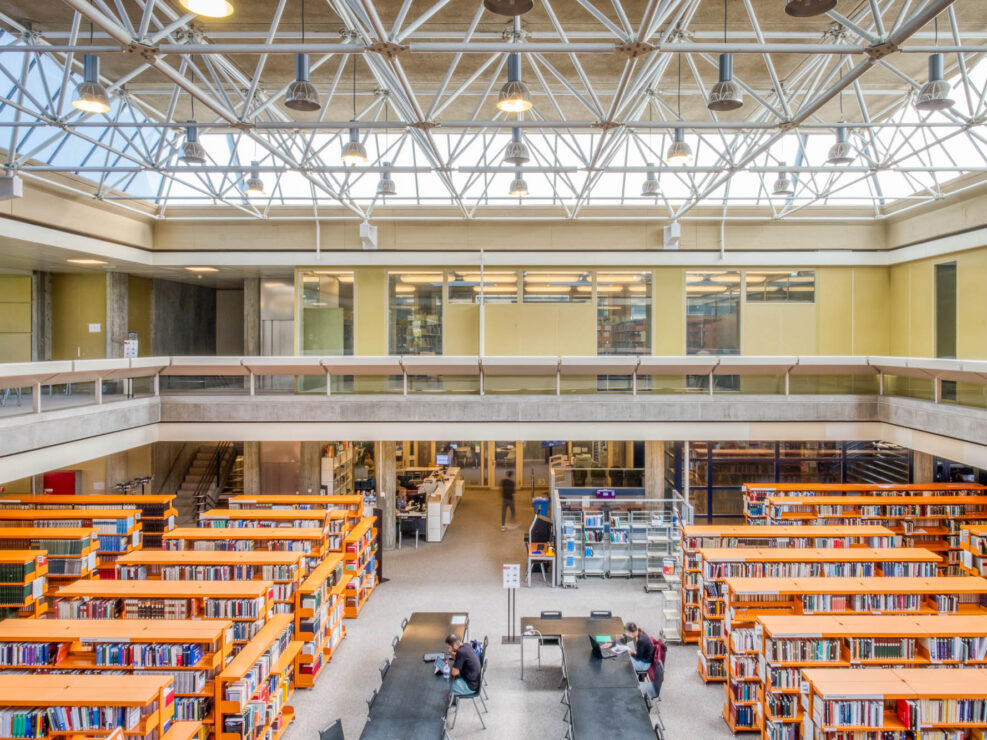 A spacious, modern library interior with tall bookshelves, tables and chairs arranged for study, a mezzanine level with glass railings, and a ceiling with exposed white beams and large windows letting in natural light.
