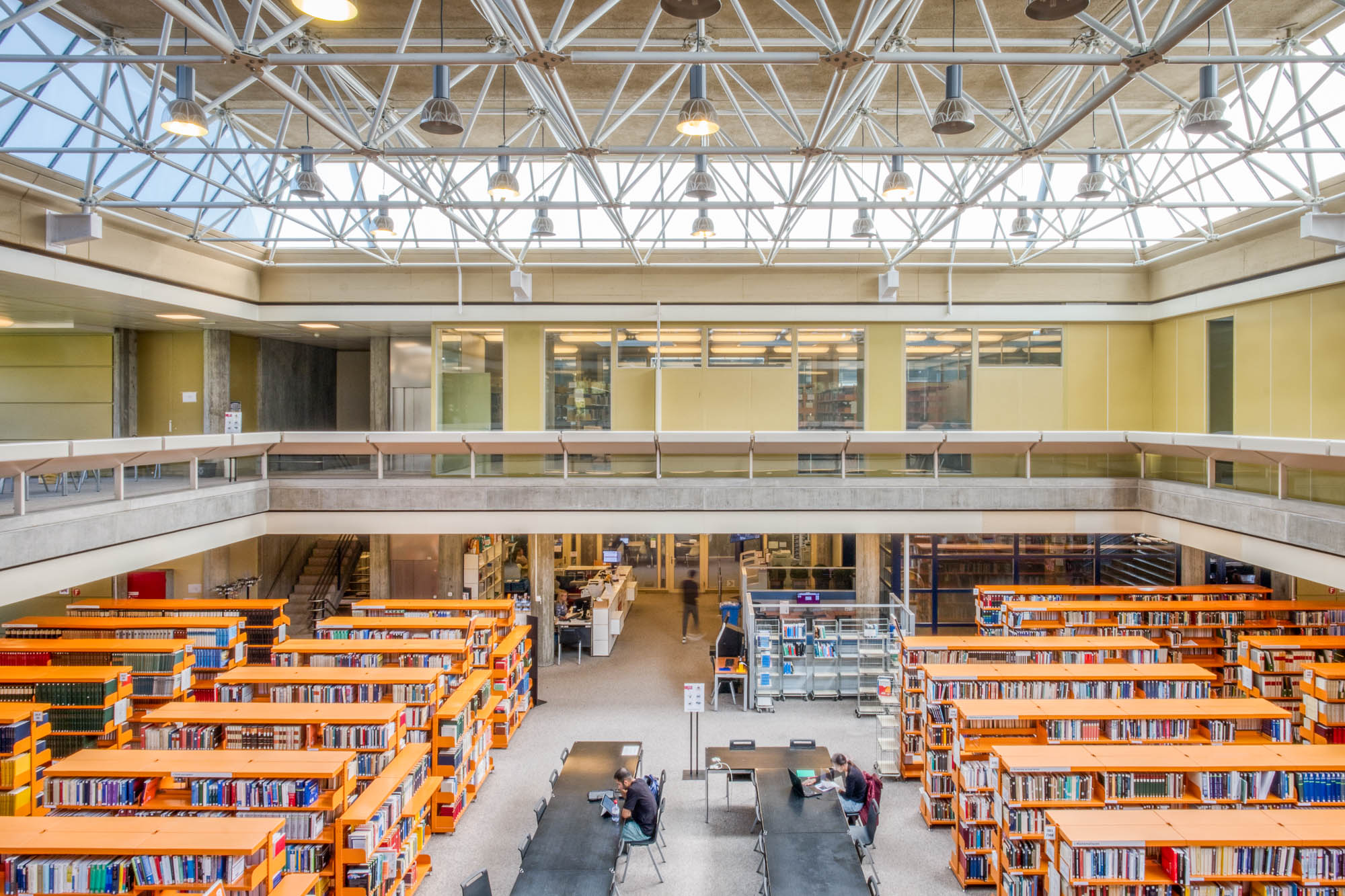 A spacious, modern library interior with tall bookshelves, tables and chairs arranged for study, a mezzanine level with glass railings, and a ceiling with exposed white beams and large windows letting in natural light.