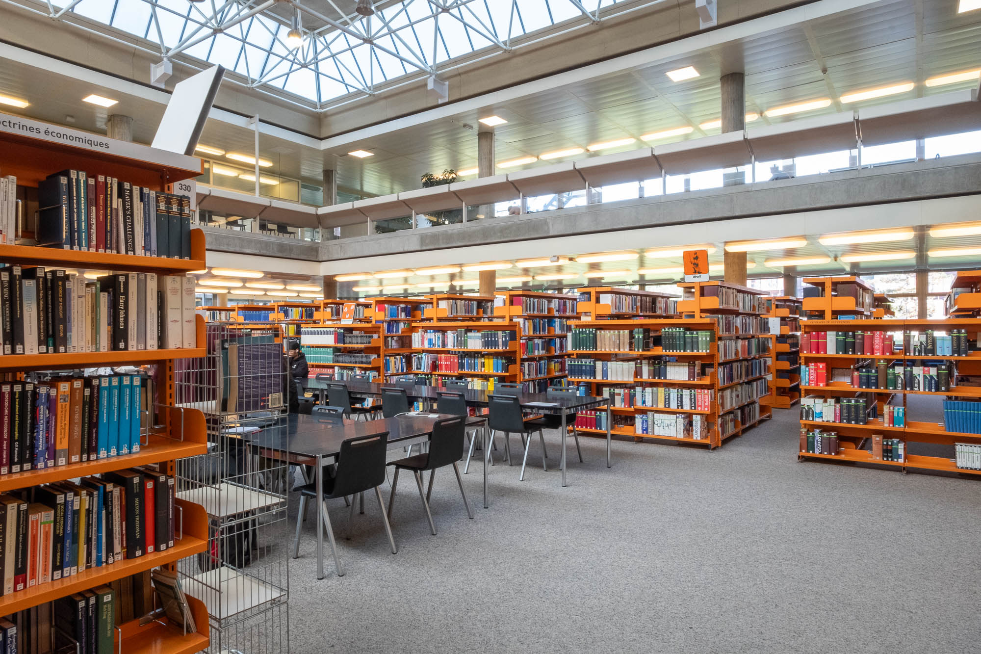 A spacious library interior with orange bookshelves filled with books, tables and black chairs arranged for reading or studying, and large windows letting in natural light from the ceiling above.