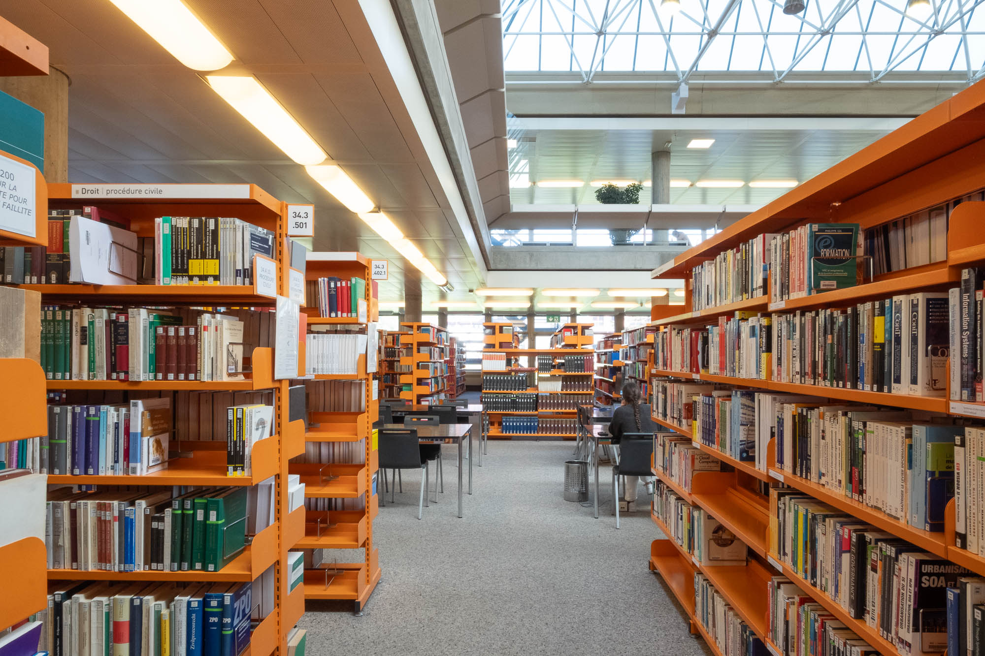 Rows of orange bookshelves filled with books line a library space with a high ceiling and large windows. A person sits at a table in the distance, reading, with more bookshelves and tables visible further back.