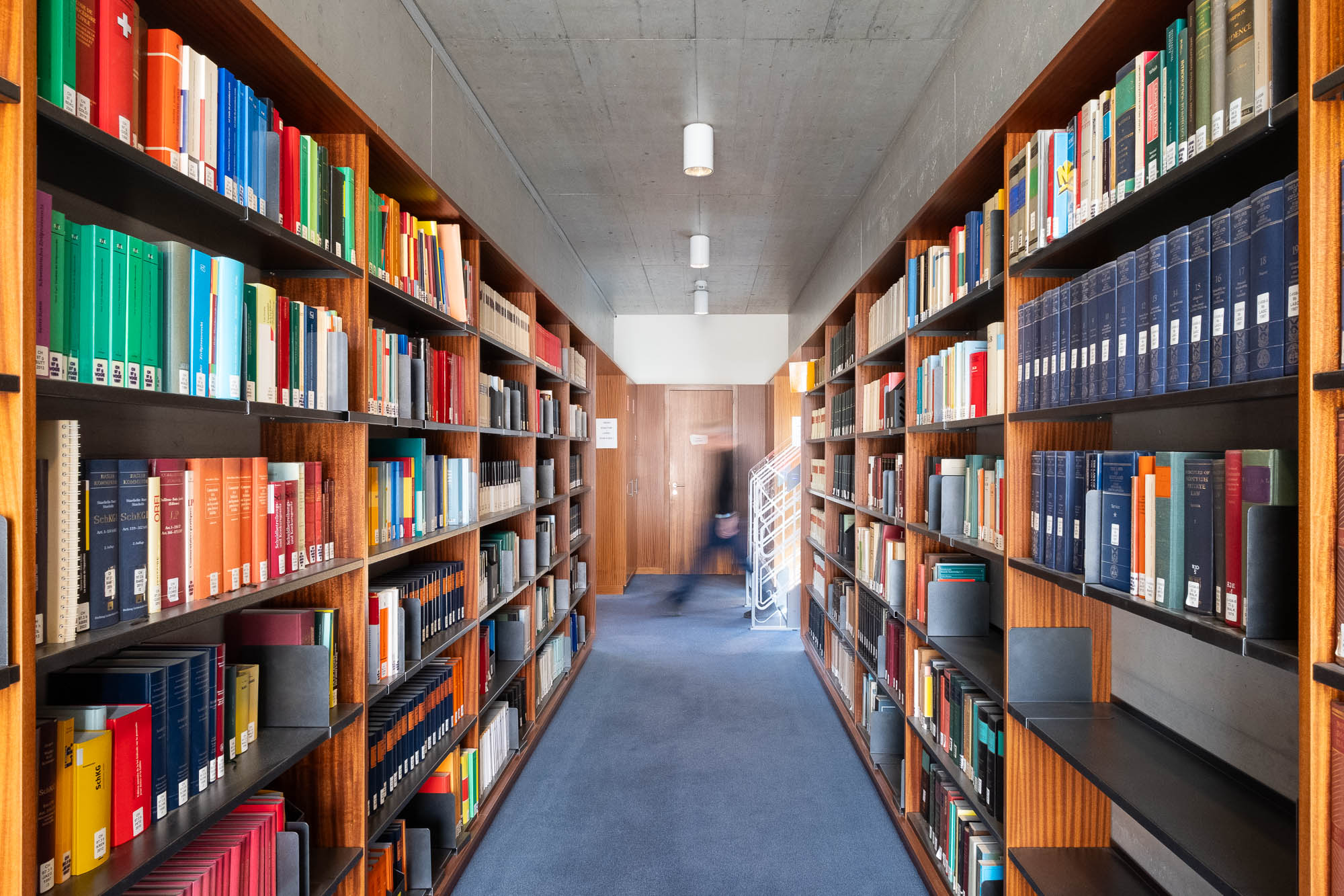 A library aisle with tall bookshelves filled with books on both sides, blue carpeted floor, and a person walking in the distance in front of a wooden wall.