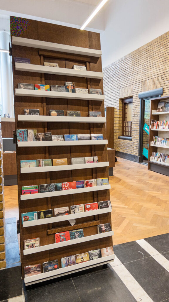 A wooden display rack in a library holds brochures and pamphlets arranged in horizontal rows, with bookshelves and a brick wall visible in the background.