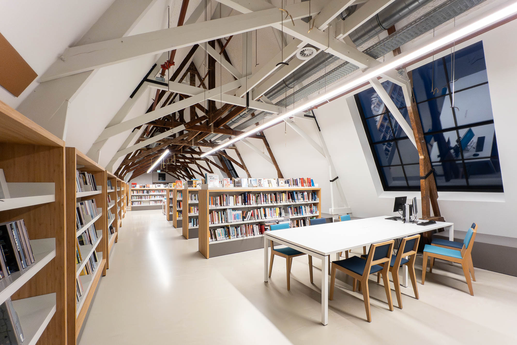 A modern library interior with high ceilings, exposed beams, bookshelves filled with books, a table with chairs, and a desktop computer near a large window. The space is well-lit with overhead lighting.