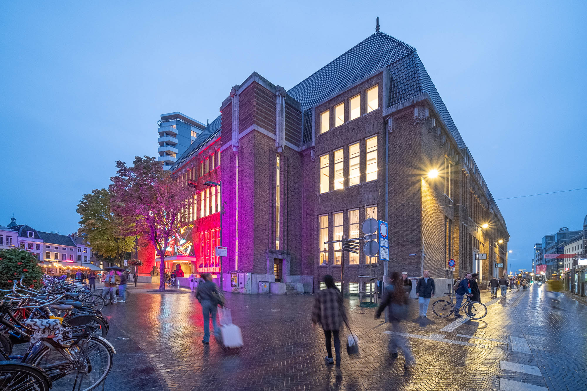A large brick building with lit windows stands on a street corner at dusk. People walk and bike past, and bicycles are parked on the sidewalk. Some trees are visible, and purple lights illuminate part of the building.