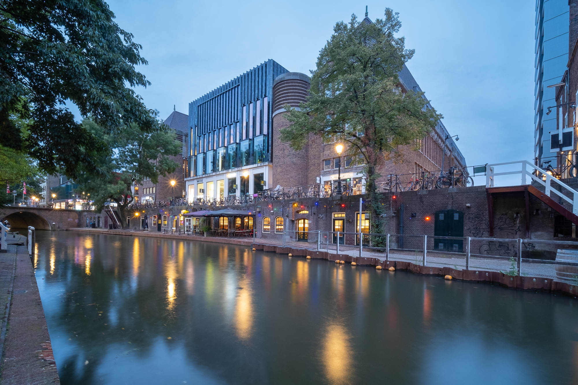 A calm canal lined with brick buildings and trees runs through a city at dusk. Lights from the buildings reflect on the water. Bicycles are parked along the edge, and a bridge crosses the canal at the back.
