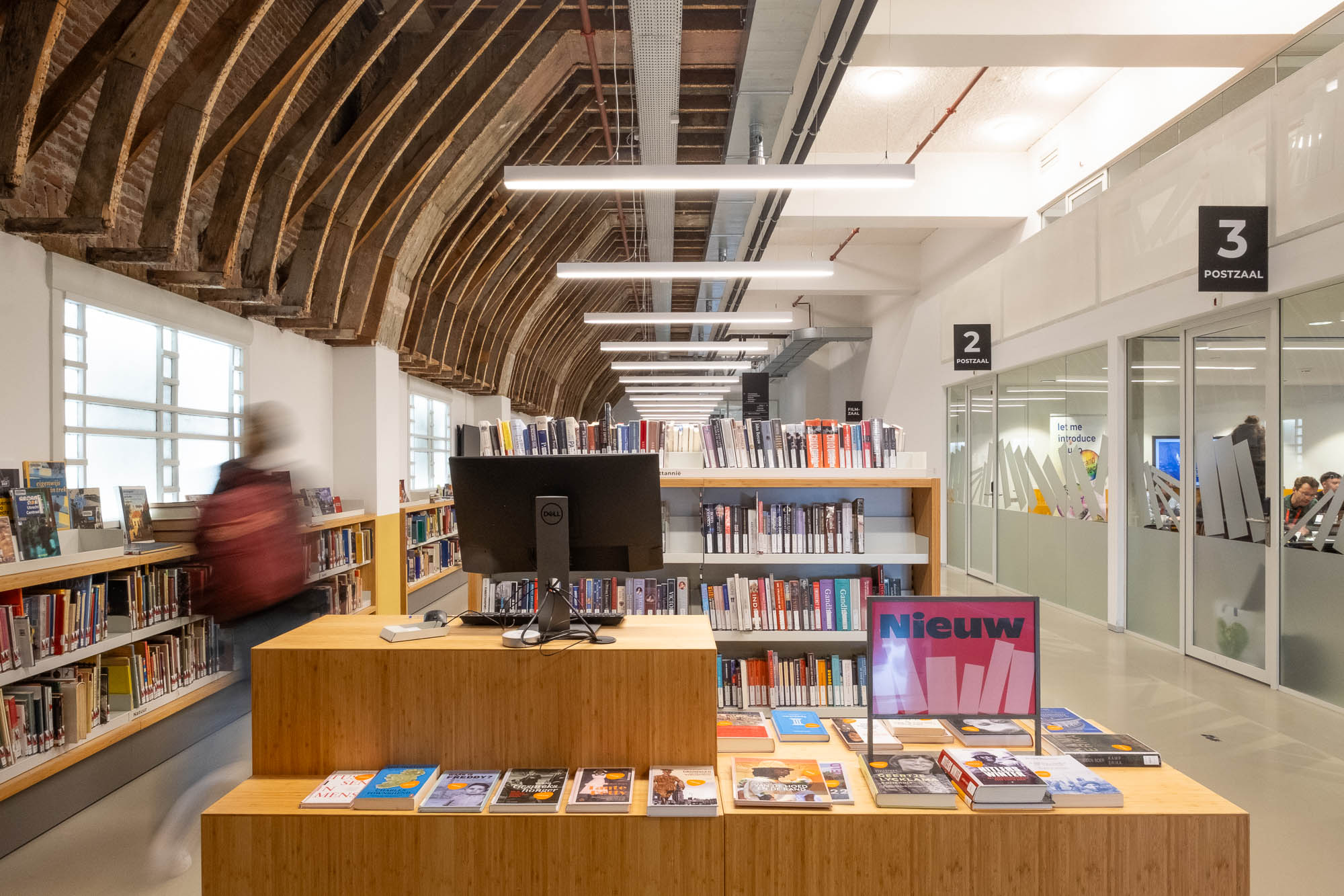 A modern library interior with wooden bookshelves, a computer on a desk, and books on display. A blurred person walks past, and there are exposed brick arches on the ceiling with glass-walled rooms on the right.