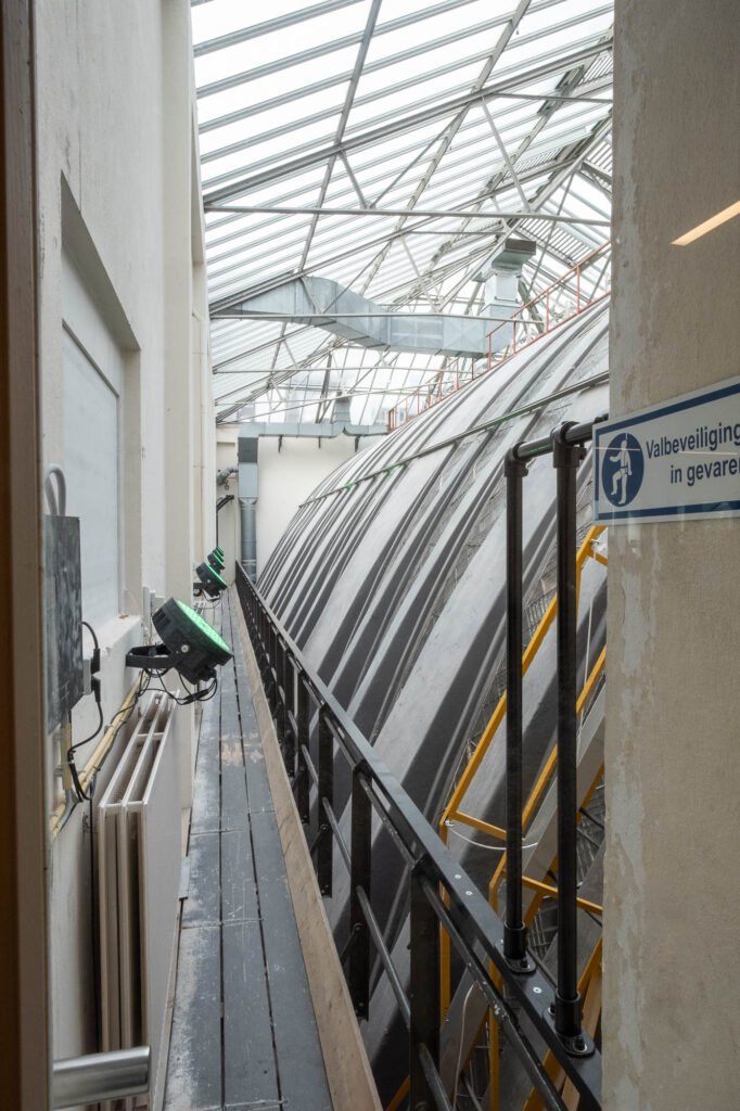 A narrow walkway with railings runs alongside a large industrial dome structure under a glass roof. There are warning lights, pipes, and a safety sign on the wall.