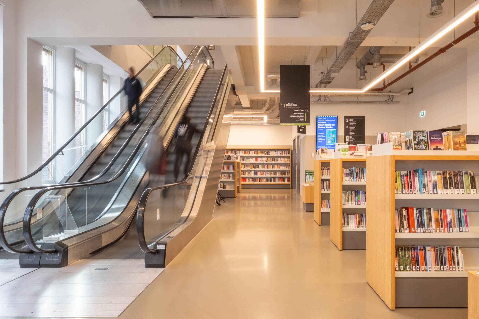 A modern library interior with bookshelves on the right, escalators on the left, and a few people going up. There are large windows, fluorescent lights, and an information screen visible in the background.