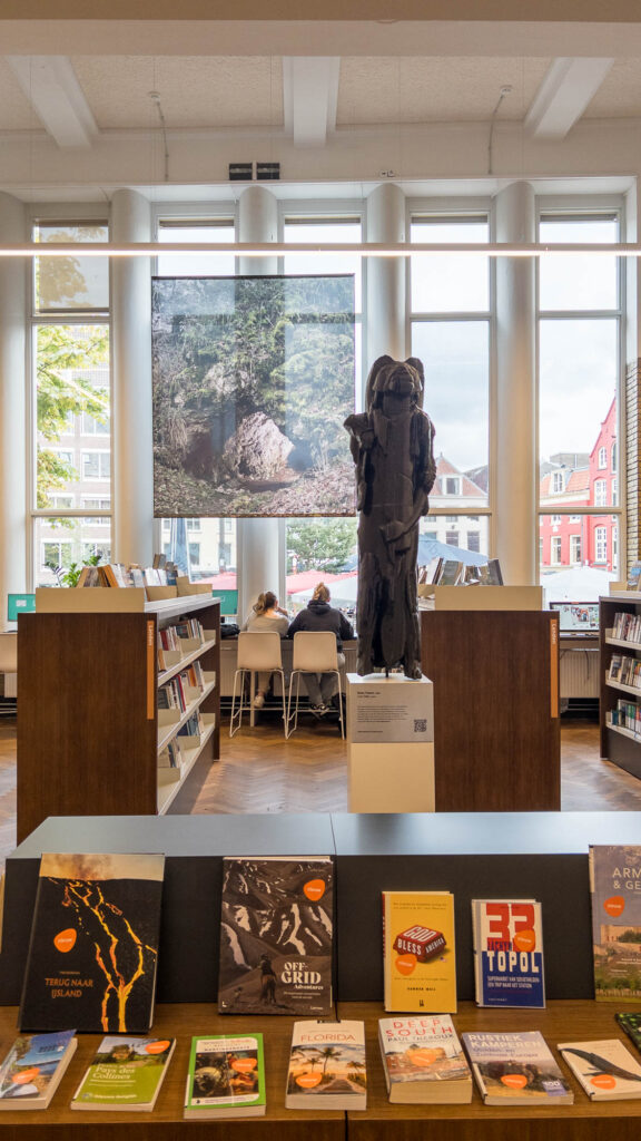 A library interior with bookshelves, a dark sculpture at the centre, and two people seated at a table by large windows. Books are displayed in the foreground; buildings and trees are visible outside.