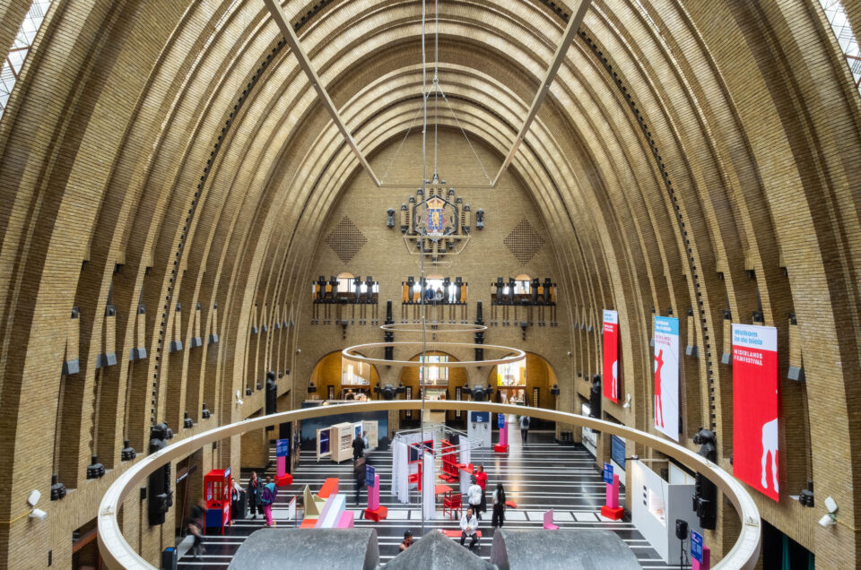An interior view of a large hall with a high arched brick ceiling, a chandelier, a circular balcony, striped flooring, display booths, and several red and white banners hanging from the balcony.