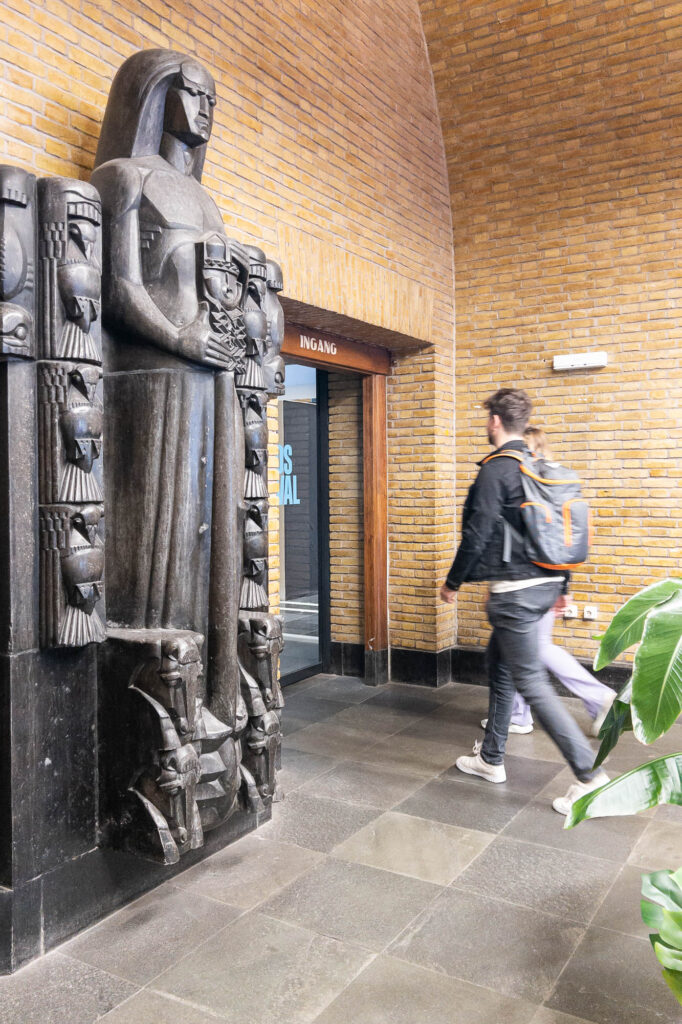 A person with a backpack walks toward a doorway labeled INGANG in a brick building. A large, dark statue of a standing person holding objects is positioned near the entrance. A plant is visible in the lower right corner.
