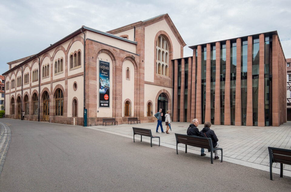 A street view of a historic building with arched windows next to a modern glass and stone structure. Three people sit on benches in the foreground while two others walk by the buildings.