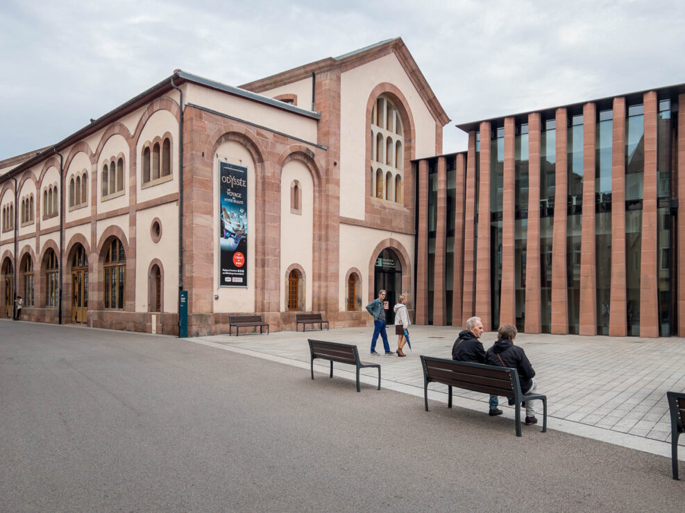 A street view of a historic building with arched windows next to a modern glass and stone structure. Three people sit on benches in the foreground while two others walk by the buildings.