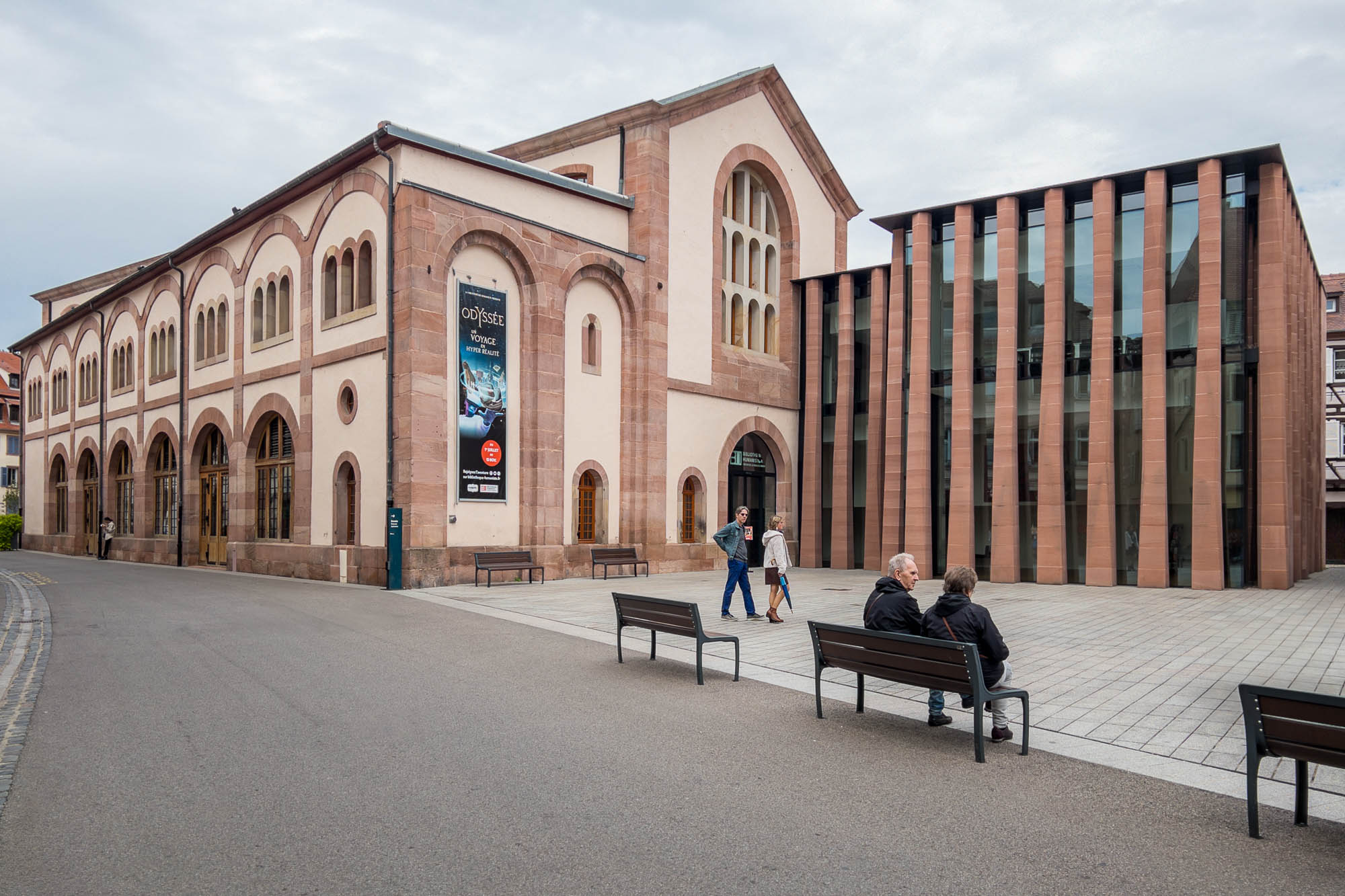 A street view of a historic building with arched windows next to a modern glass and stone structure. Three people sit on benches in the foreground while two others walk by the buildings.