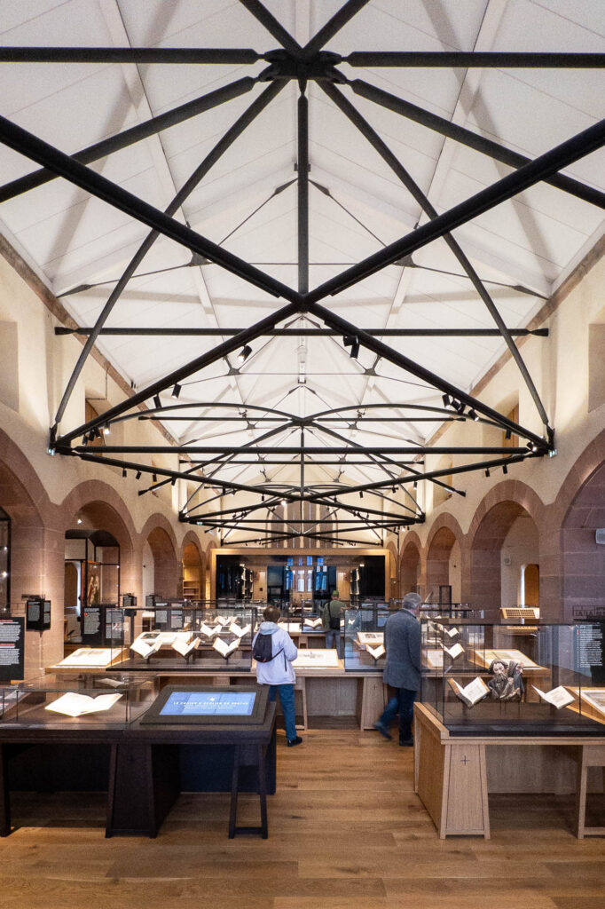 People are strolling between display cases in an exhibition space with wooden floors, bookshelves, and modern glass partitions under a high, vaulted ceiling with metal beams.