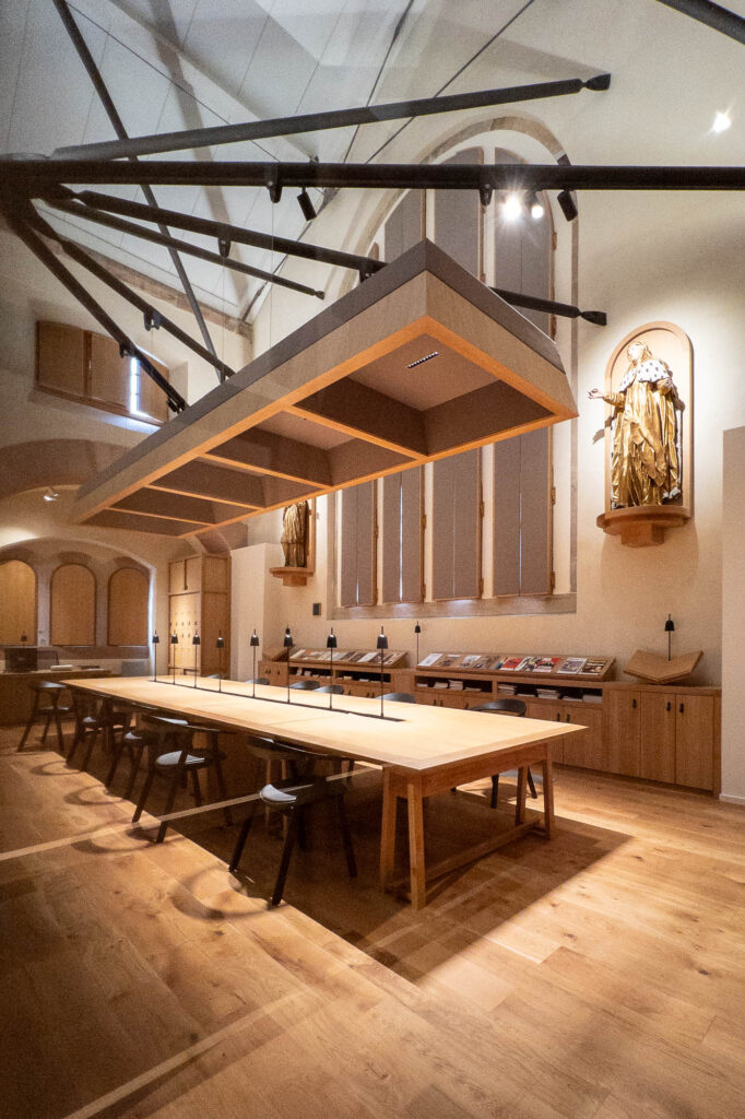 A spacious, well-lit room features a long wooden table with black chairs, overhead rectangular lighting, statues in wall niches, bookshelves along the wall, and arched windows with vertical blinds.