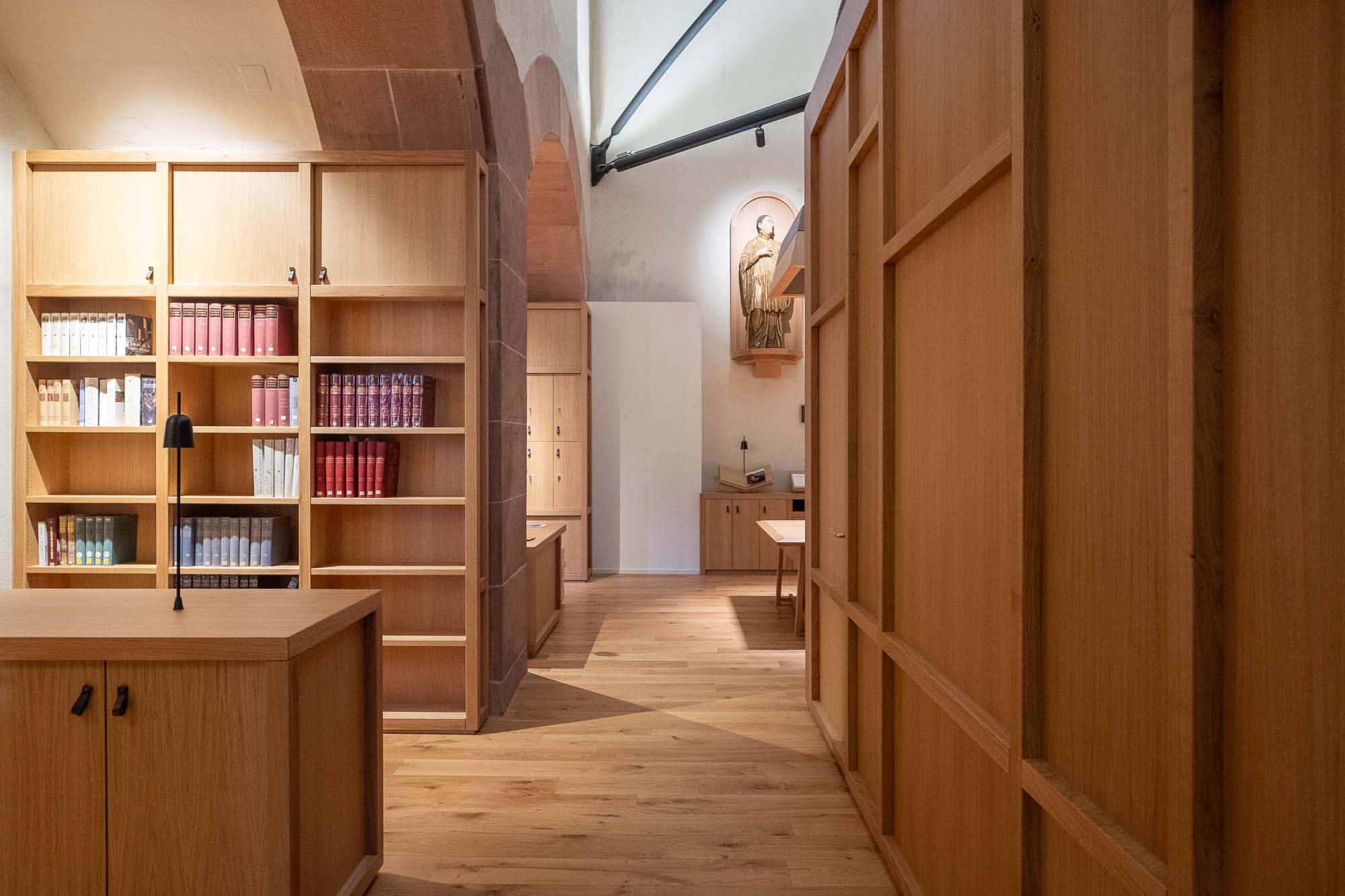 A library or study room with wooden bookshelves and cabinets, a lamp on a desk, books arranged on the shelves, and a religious statue displayed in a niche on the far wall.