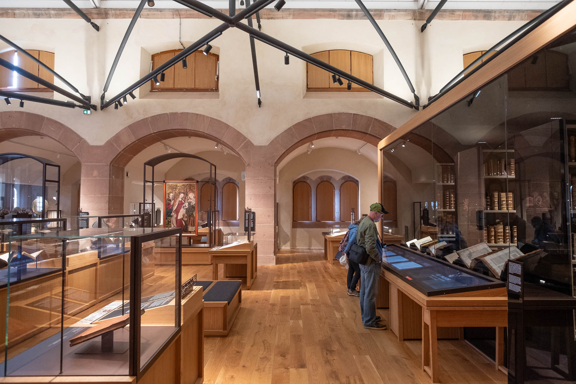Two people stand side by side, reading a display case in an exhibition space with wooden floors, bookshelves, and modern glass partitions under a high, vaulted ceiling with metal beams.