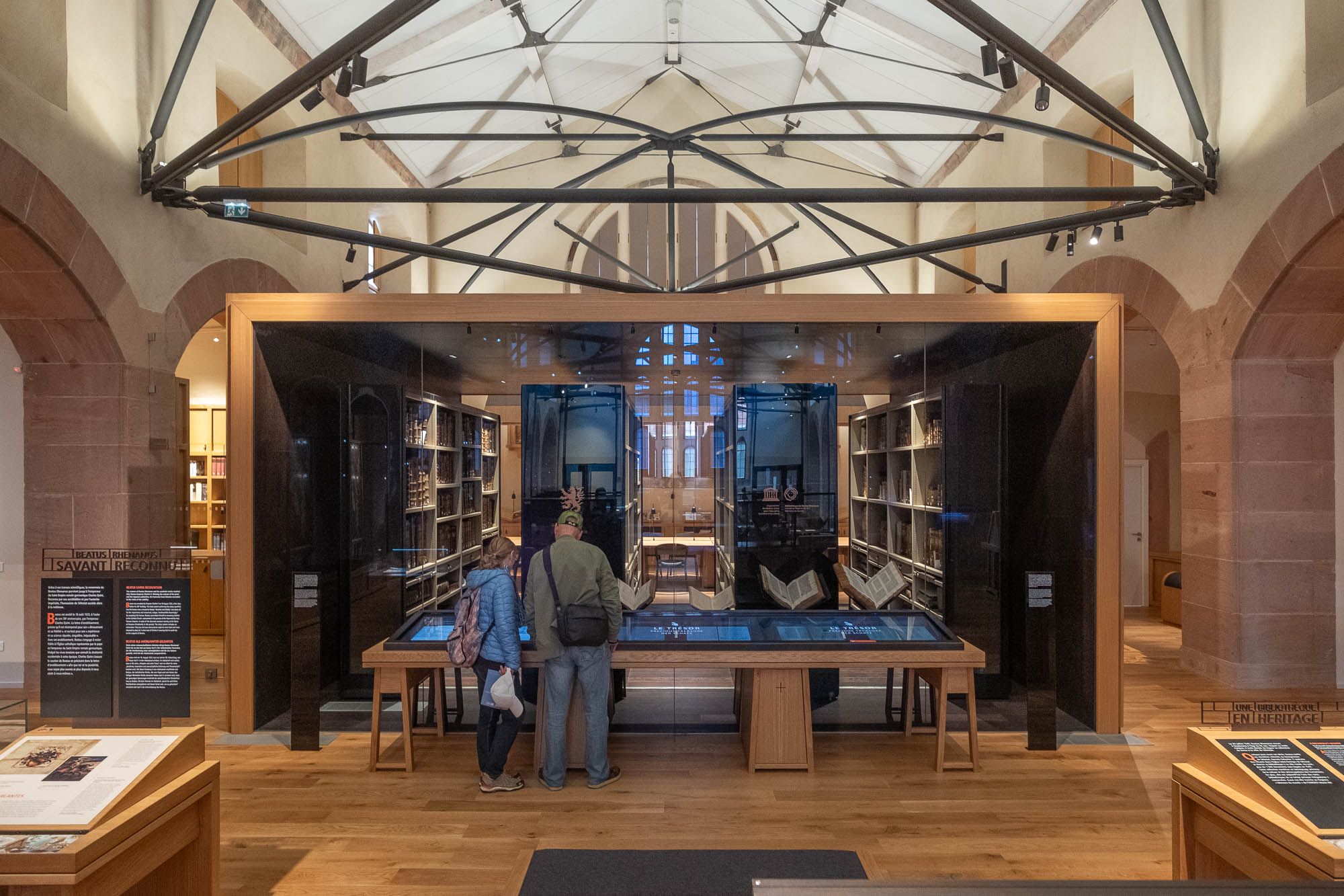 Two people stand side by side, reading a display case in an exhibition space with wooden floors, bookshelves, and modern glass partitions under a high, vaulted ceiling with metal beams.