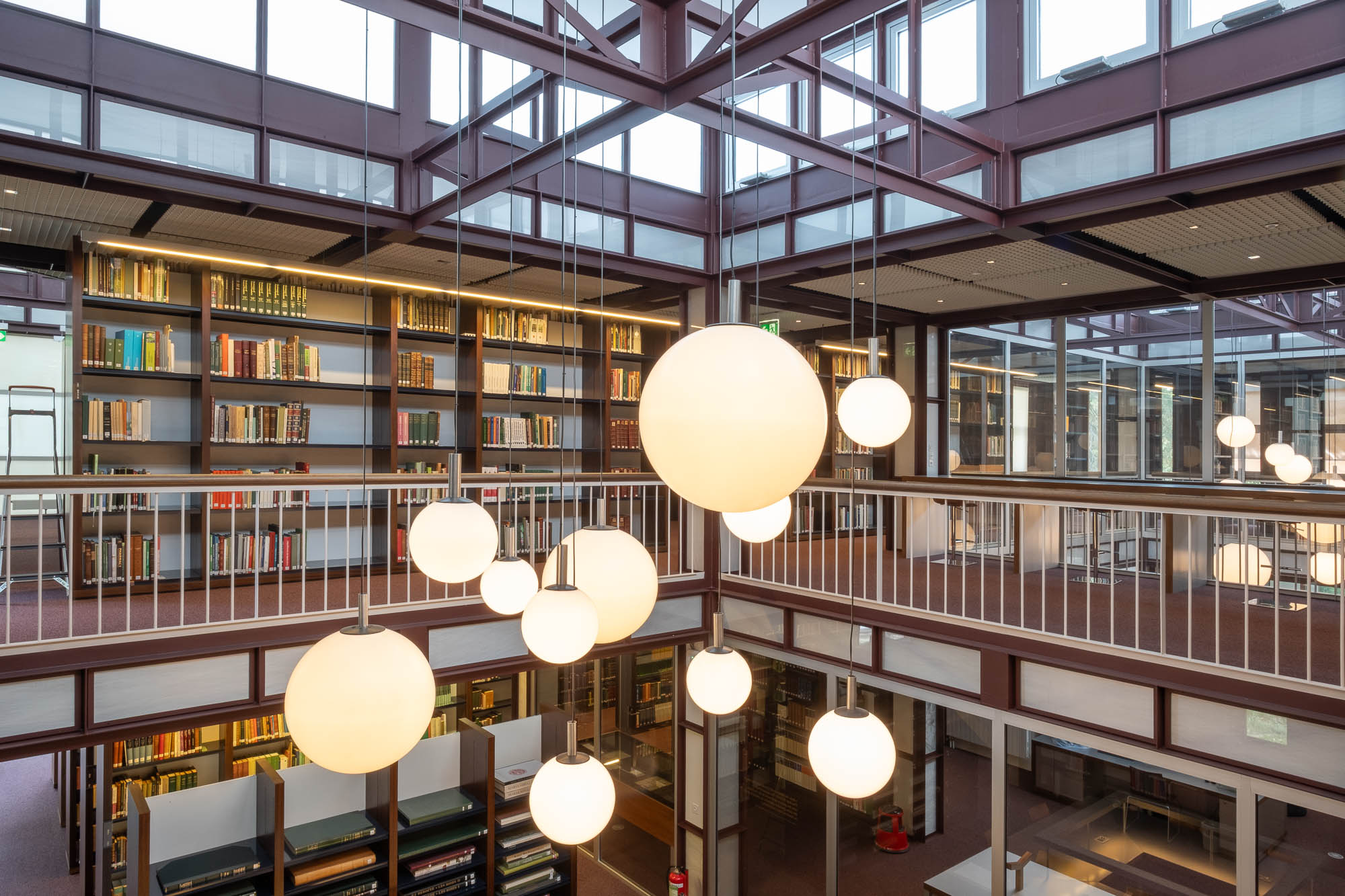 Large round light fixtures hang from the ceiling in a modern two-story library, which features rows of bookshelves, glass walls, and an open balcony overlooking the lower level.