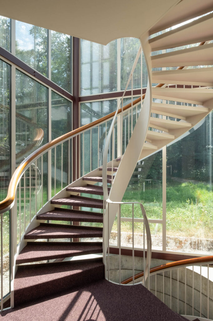 A modern spiral staircase with white railings and wooden handrails, set next to large glass windows with views of green trees and grass outside. Natural light fills the stairwell.