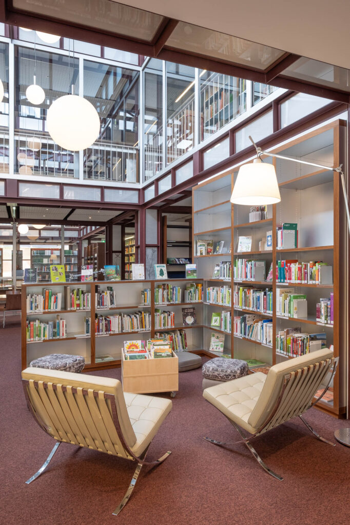 Modern library interior with shelves of books, two cream-coloured chairs, a small table, and a floor lamp. Large windows and glass walls let natural light into the space.
