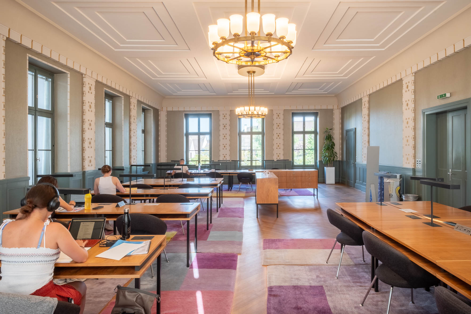 Spacious, well-lit room with several people seated at large wooden tables working on laptops. The room features tall windows, modern chandeliers, and a colorful patterned rug.