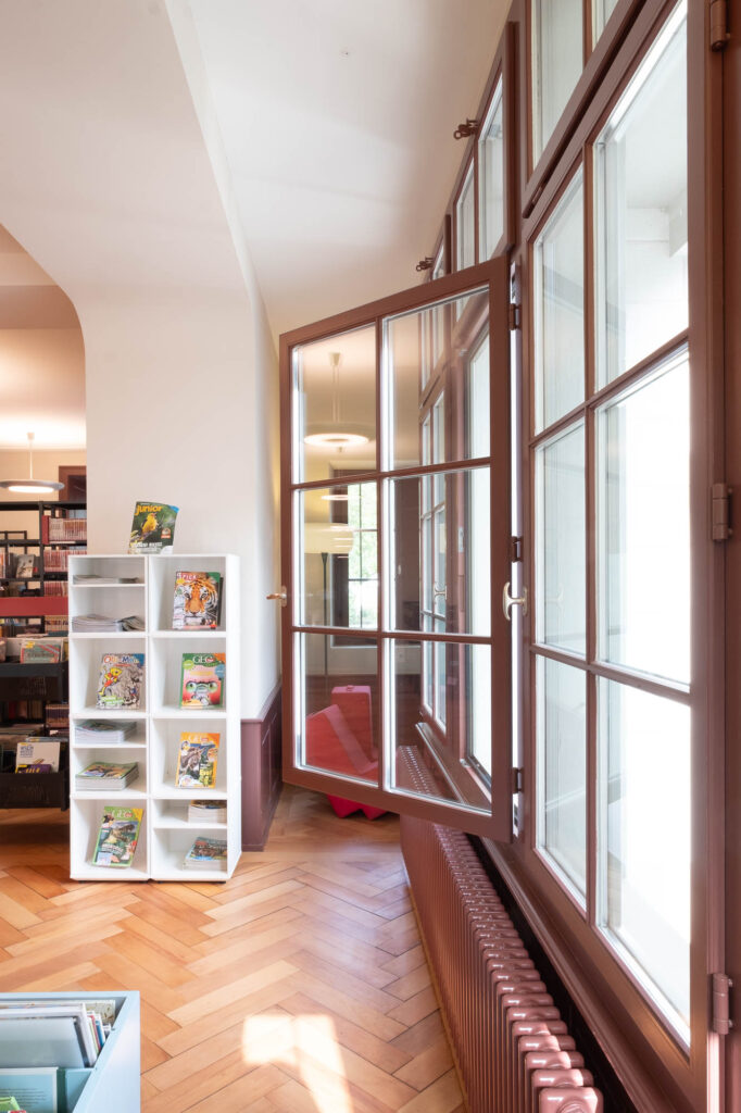 A large window is open in a room with wooden herringbone flooring. To the left, a white shelf displays colorful magazines and books. There is a partial view of additional shelves and lighting in the background.
