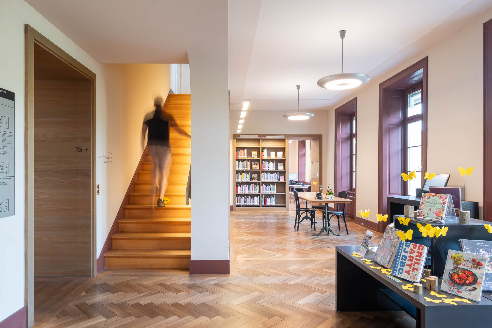 A person ascends a wooden staircase in a brightly lit library space with bookshelves, tables, and a display rack decorated with yellow butterfly cutouts. The wooden floor has a herringbone design.