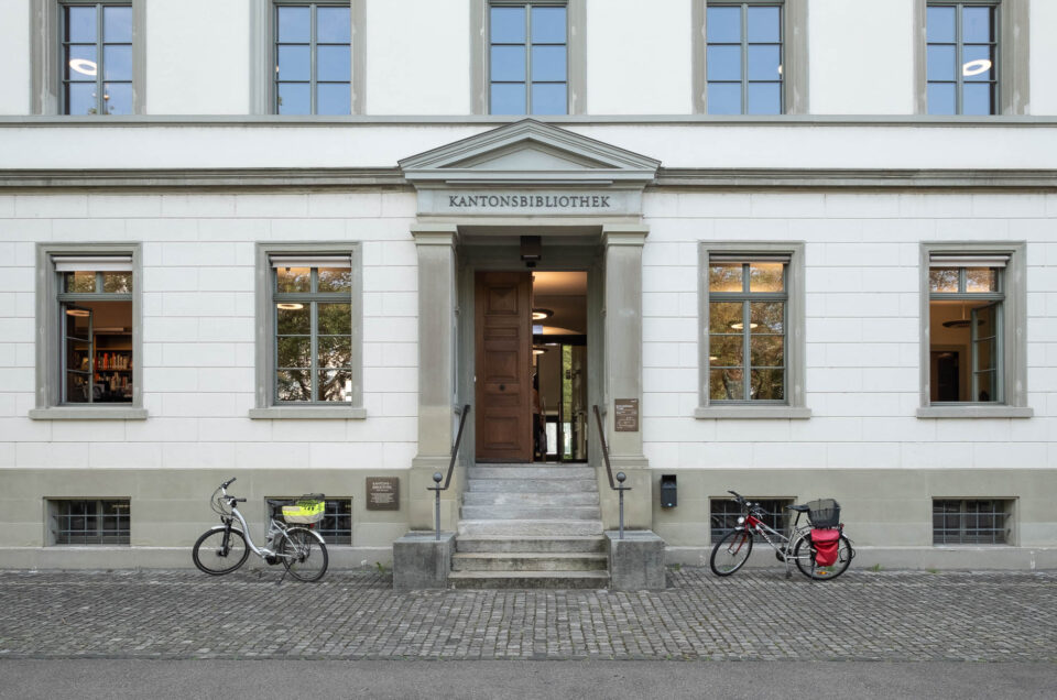 The entrance to a white building labeled Kantonsbibliothek with a wooden door under a portico. Bicycles are parked on either side of the entrance.