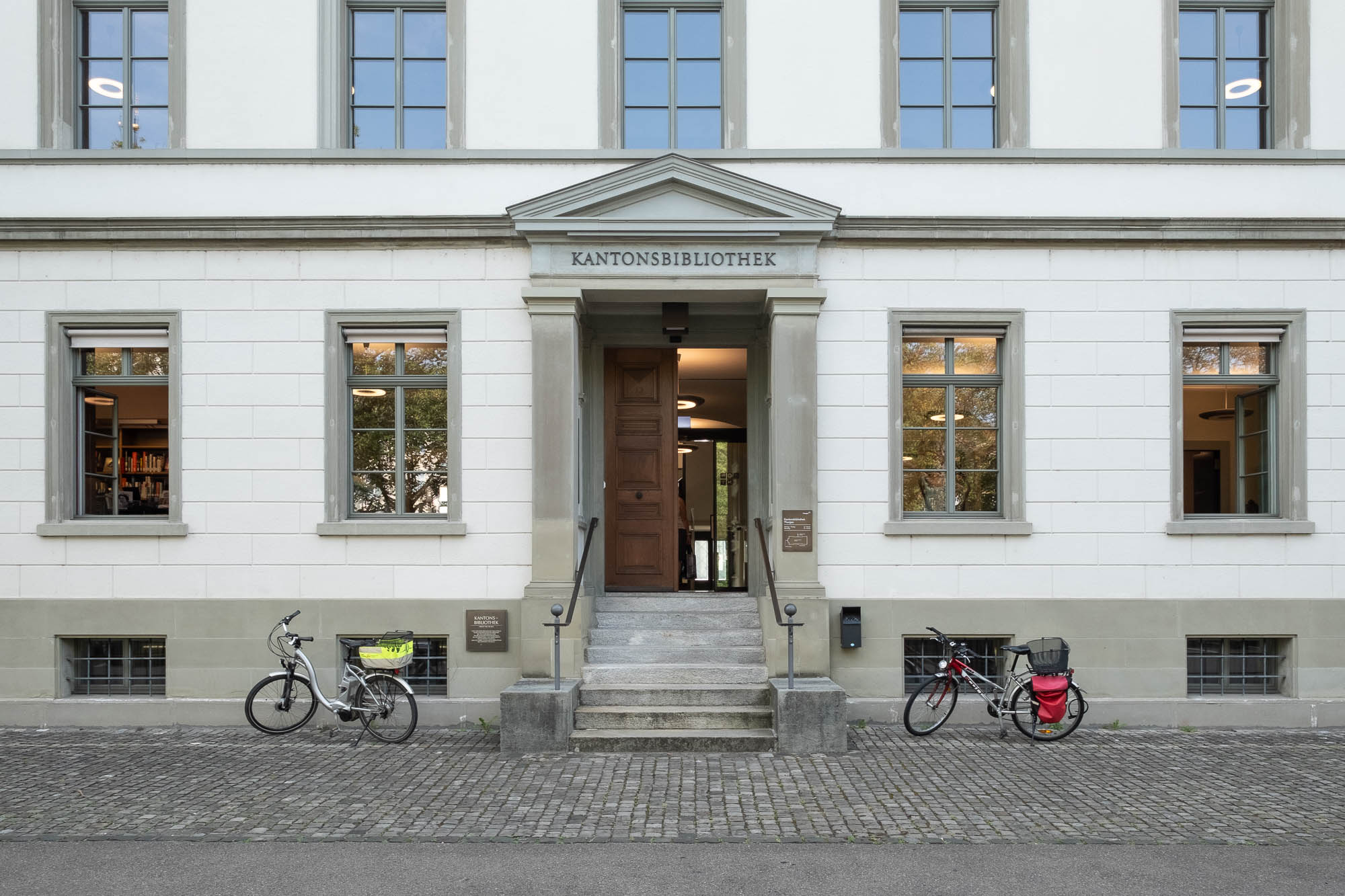The entrance to a white building labeled Kantonsbibliothek with a wooden door under a portico. Bicycles are parked on either side of the entrance.